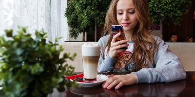 young girl sits at cafe table with drink while looking at her cell phone
