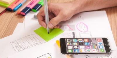 photo of desk with papers and person marking on a sticky note with their phone open to apps