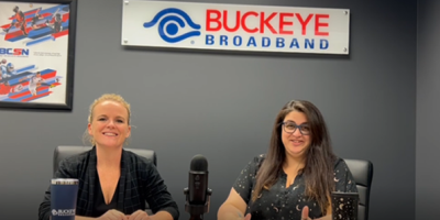 kim, blonde and smiling, sits next to rani, brunette and happy, under the buckeye broadband logo with a mic on the table