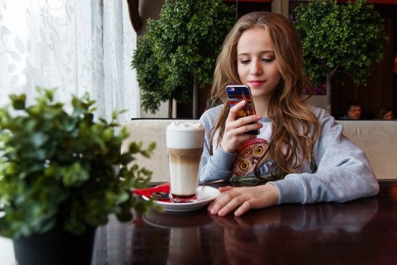 young girl sits at cafe table with drink while looking at her cell phone