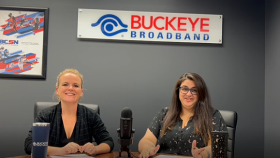 kim, blonde and smiling, sits next to rani, brunette and happy, under the buckeye broadband logo with a mic on the table