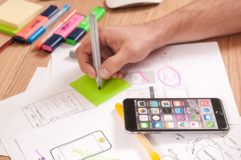 photo of desk with papers and person marking on a sticky note with their phone open to apps